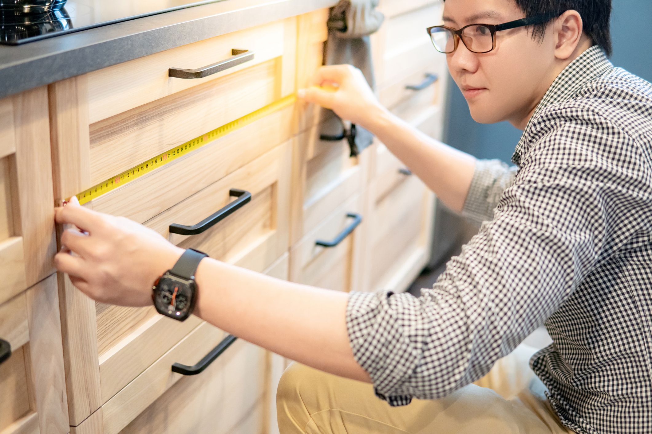 Young Asian man using tape measure for measuring drawer on wooden kitchen counter in showroom. Shopping furniture for home improvement. Interior design concept
