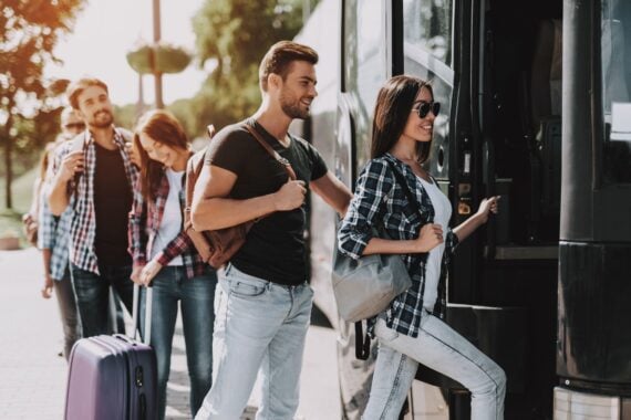 A smiling woman in a plaid shirt boards a parked tour bus, followed by a man with a backpack and a line of other travelers.