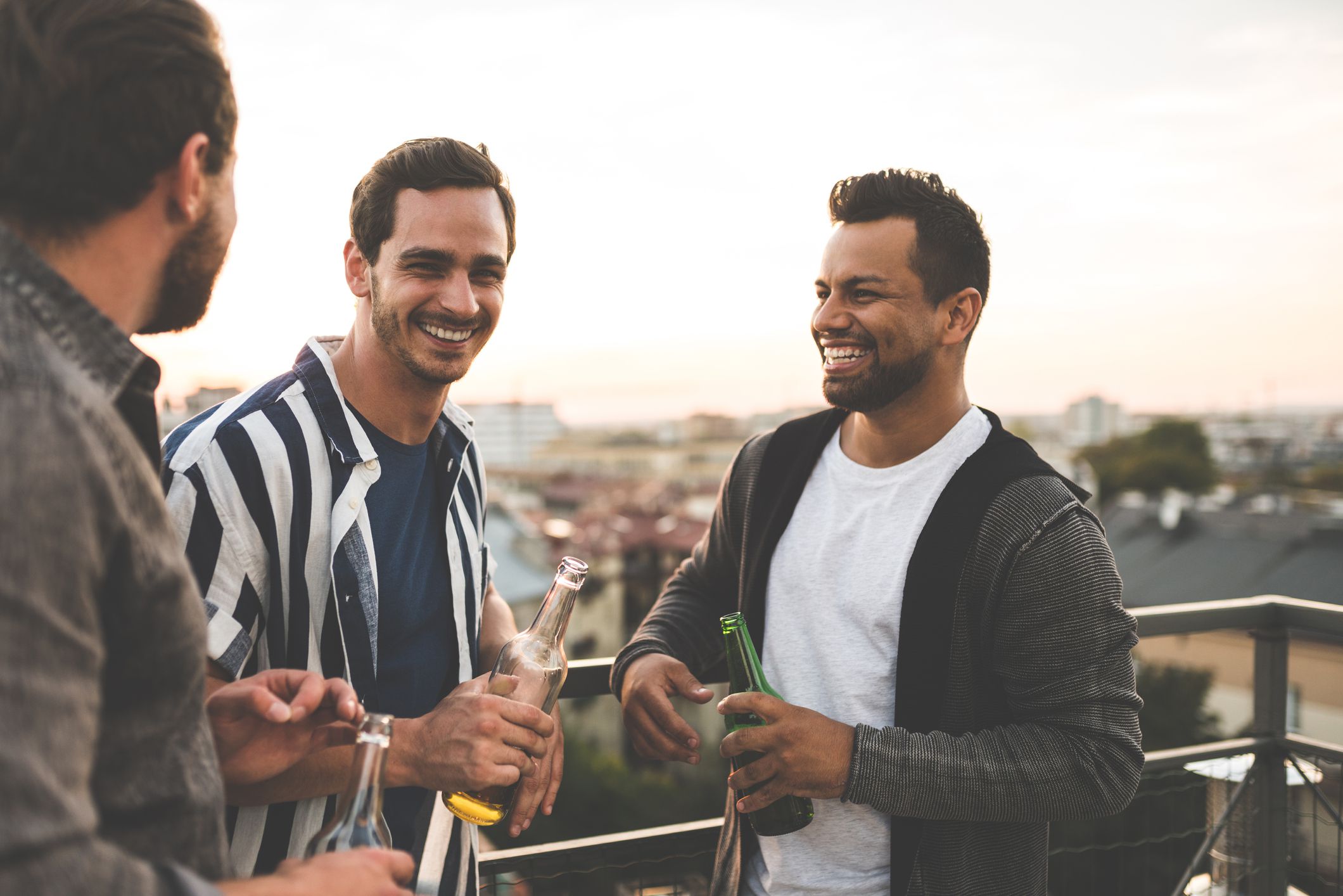 Group of male friends standing together at rooftop with drinks and chatting. Cheerful friends enjoying at terrace party.