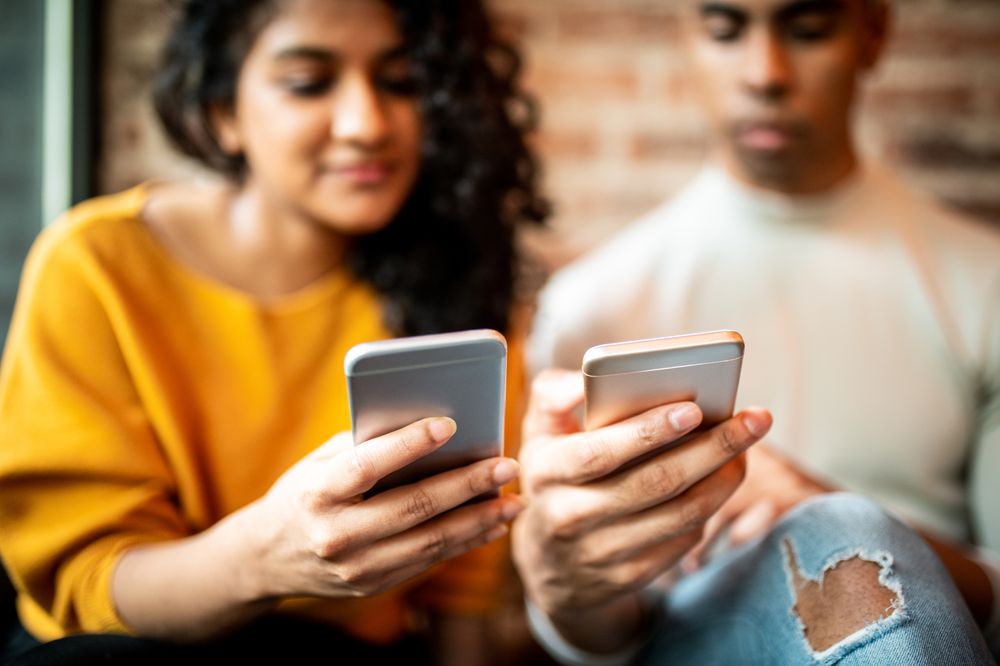 Two people sitting closely together, both holding and looking at their smartphones. The person on the left is wearing a yellow sweater, and the person on the right is wearing a light-colored shirt and ripped jeans.