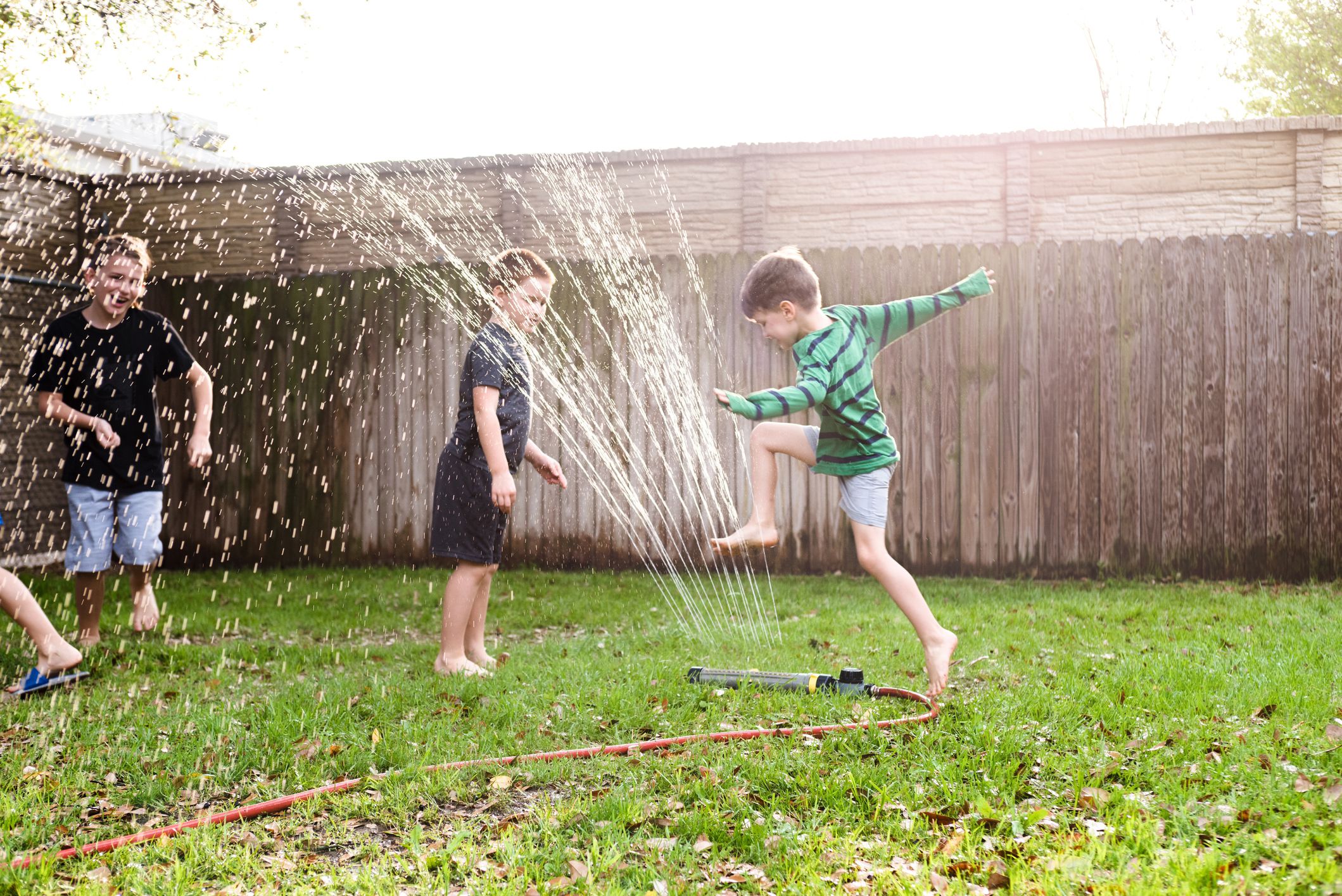 Brothers run and jump over the sprinkler to cool off on a hot summer day. They are playing in their backyard.