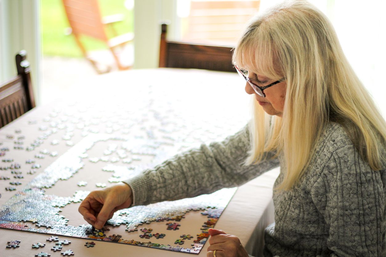 Color image depicting a senior caucasian couple, in their 60s and 70s, doing a jigsaw puzzle together at home during the coronavirus (Covid-19) pandemic. The couple are both wearing spectacles and are concentrating on completing the puzzle together.