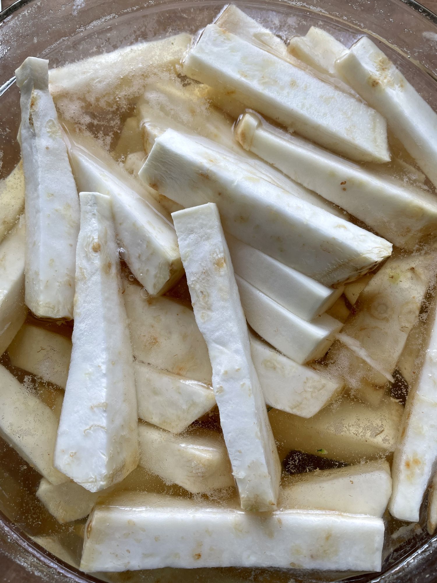Stock photo showing an elevated view of a wooden chopping board containing a glass bowl full of sliced celeriac wedges soaking in water.
