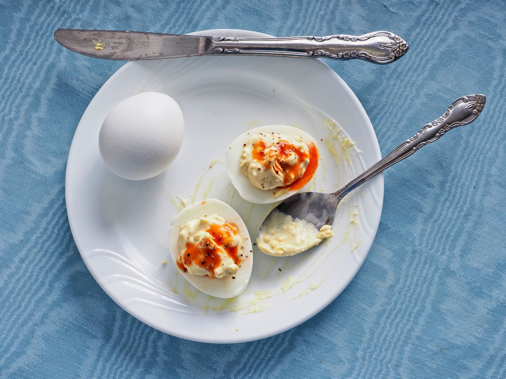Eggs of the devil, spicy deviled eggs with red hot sauce on top a savory snack or appetizer. Prepared deviled eggs on a white plate with blue tablecloth background.