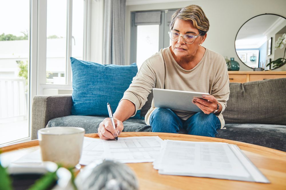 Shot of a mature woman using a digital tablet while going through paperwork at home