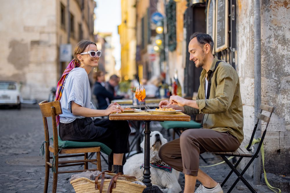 A woman and man sit at an outdoor café table in a charming, narrow street, smiling and eating with drinks on the table. A dog lies beneath the table, and a woven bag rests nearby. The atmosphere is relaxed and lively.