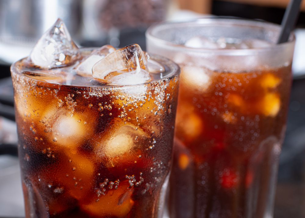 Two glasses filled with iced cola, topped with ice cubes and bubbles, sit on a table. Condensation forms on the outside of the glasses, indicating the drinks are cold.