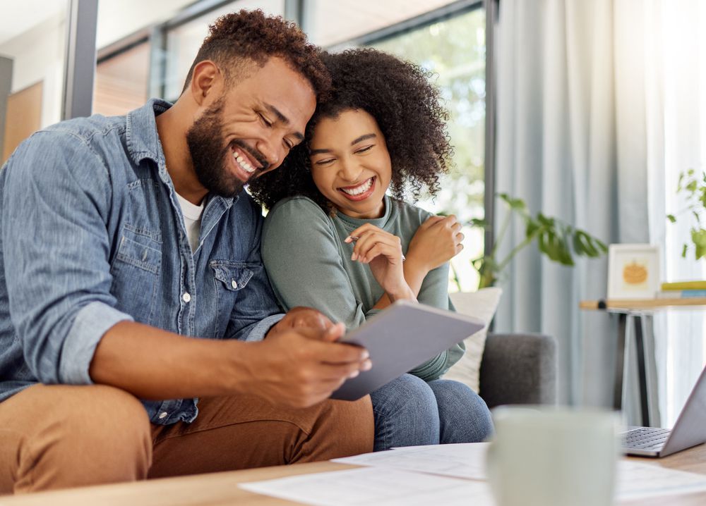 A smiling couple sits close together on a couch, looking at a tablet. Papers and a laptop are on the table in front of them, and natural light fills the cozy, modern living room.