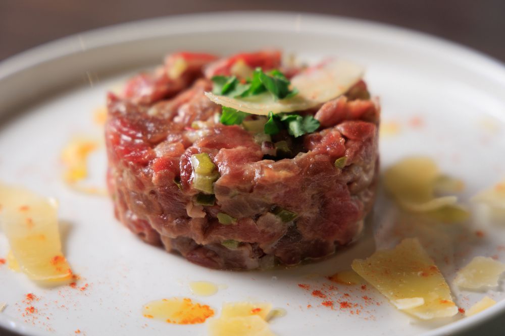 A close-up of steak tartare, garnished with chopped herbs and thin cheese slices on a white plate, with a drizzle of oil and a sprinkle of spice around the dish.