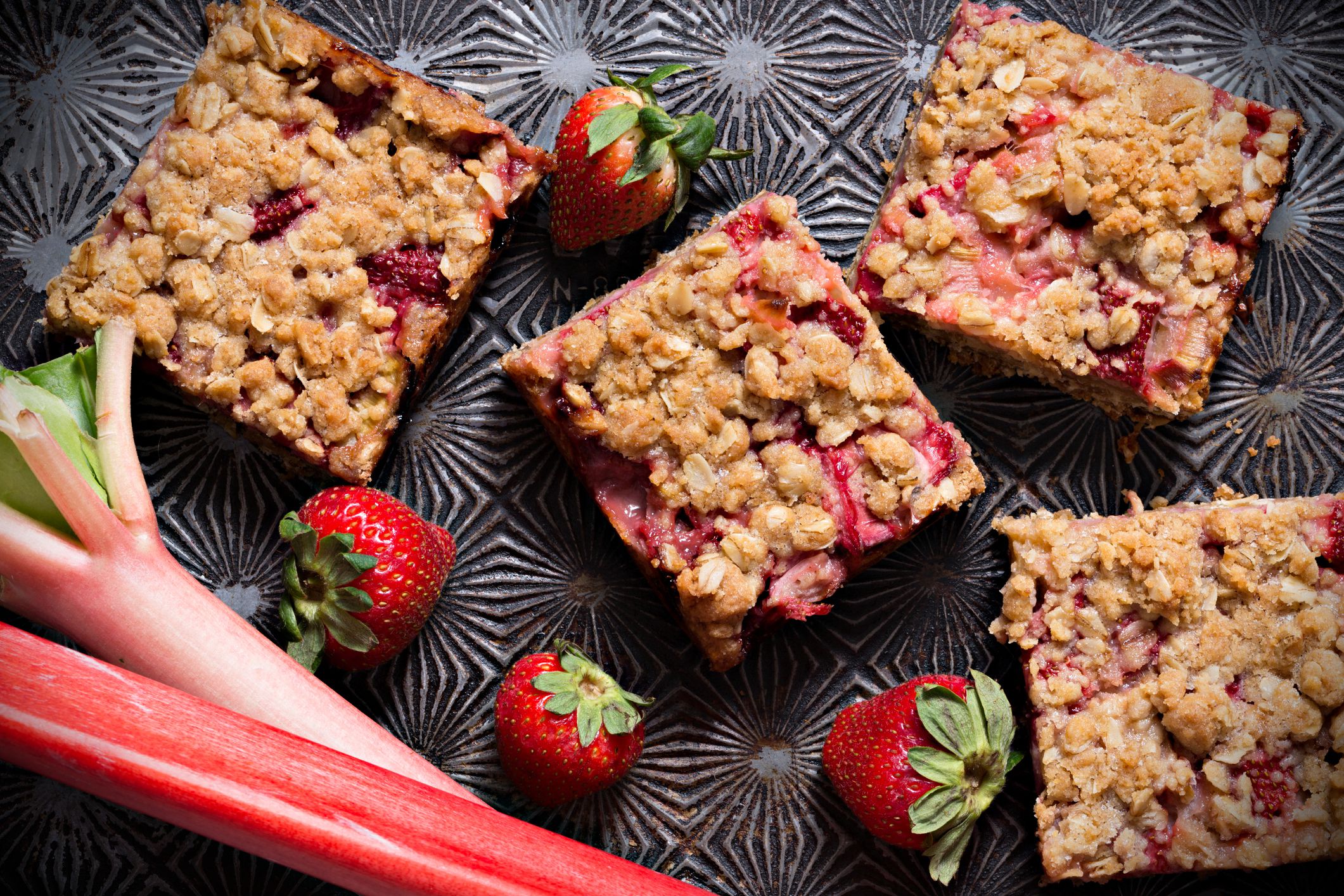 An overhead close up horizontal photograph of several homemade strawberry rhubarb bars, some fresh ripe strawberries and a couple of rhubarb stems on an old baking tray.