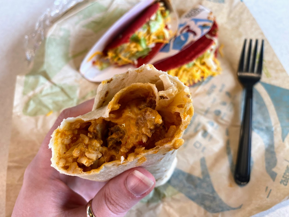 A hand holds a half-eaten burrito filled with rice, cheese, and beef. In the background, a taco with a red shell, shredded lettuce, and cheese sits on a wrapper next to a black plastic fork.