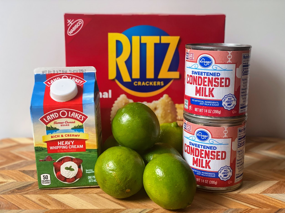 A box of Ritz crackers, a carton of Land O’Lakes heavy whipping cream, two cans of Kroger sweetened condensed milk, and four limes are arranged on a wooden surface.