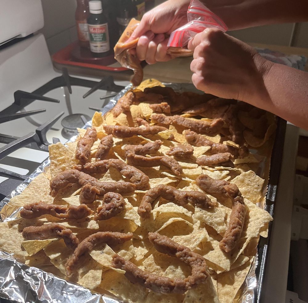 Hands squeezing refried beans from a plastic bag onto tortilla chips arranged on a foil-lined baking sheet, preparing nachos in a home kitchen.