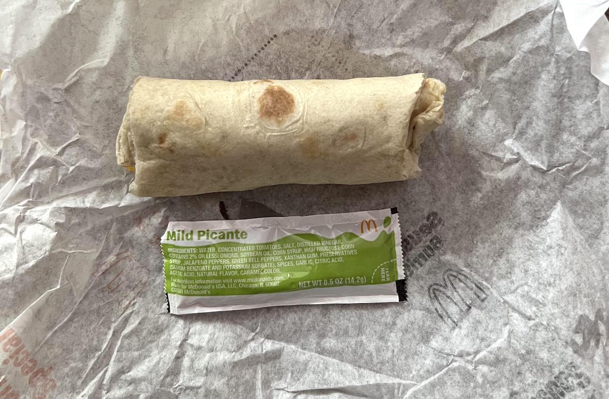 A wrapped burrito, rumored to be one of the best fast-food breakfast burritos, sits on crumpled wax paper next to an unopened packet of McDonald's Mild Picante sauce.
