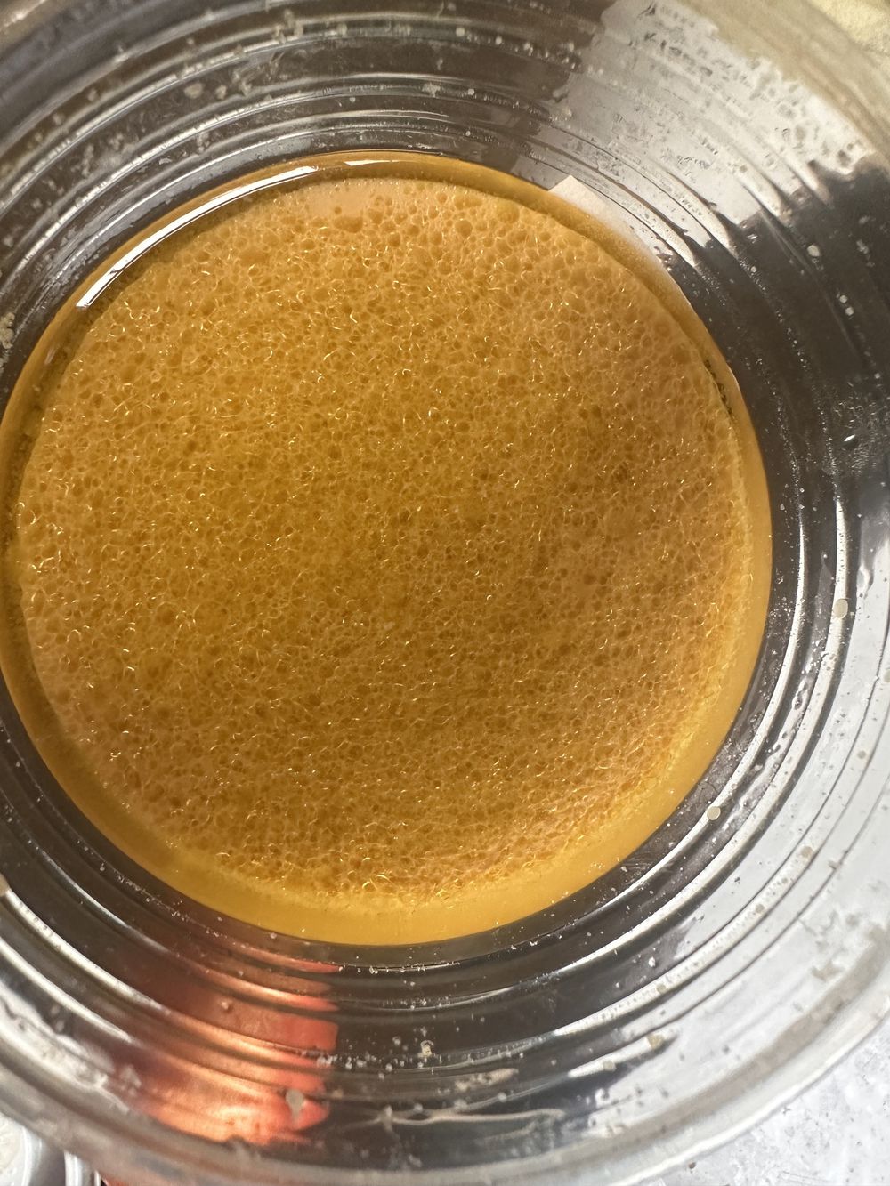 A close-up view of a glass bowl containing a light brown, frothy liquid with a layer of bubbles on the surface. The bowl is sitting on a metal surface.
