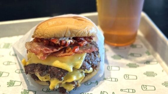 A cheeseburger with two beef patties, melted cheese, bacon, and diced red peppers sits on a tray next to a clear cup filled with a light amber drink.