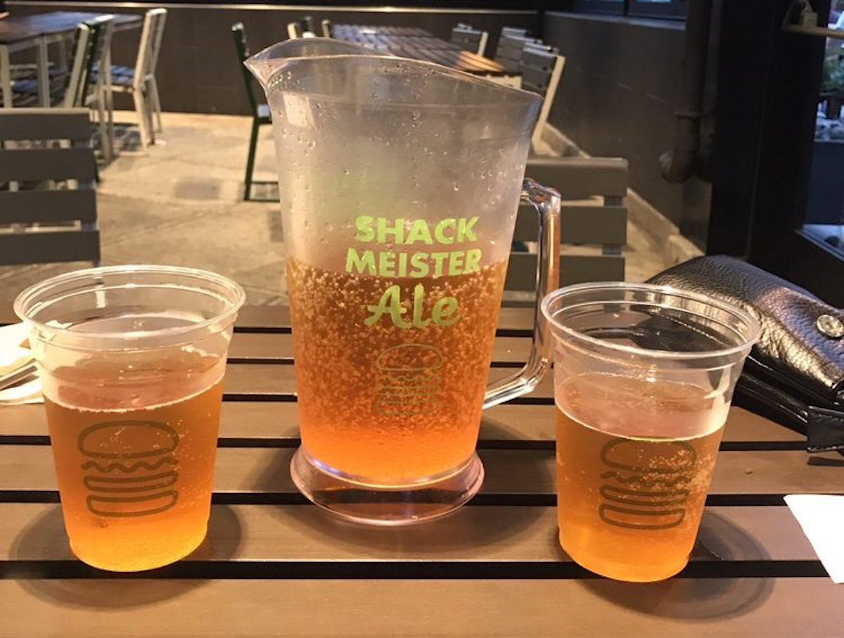 A plastic pitcher labeled "Shack Meister Ale" filled with beer sits on a table between two clear plastic cups of beer. A black wallet and chairs are visible in the background.