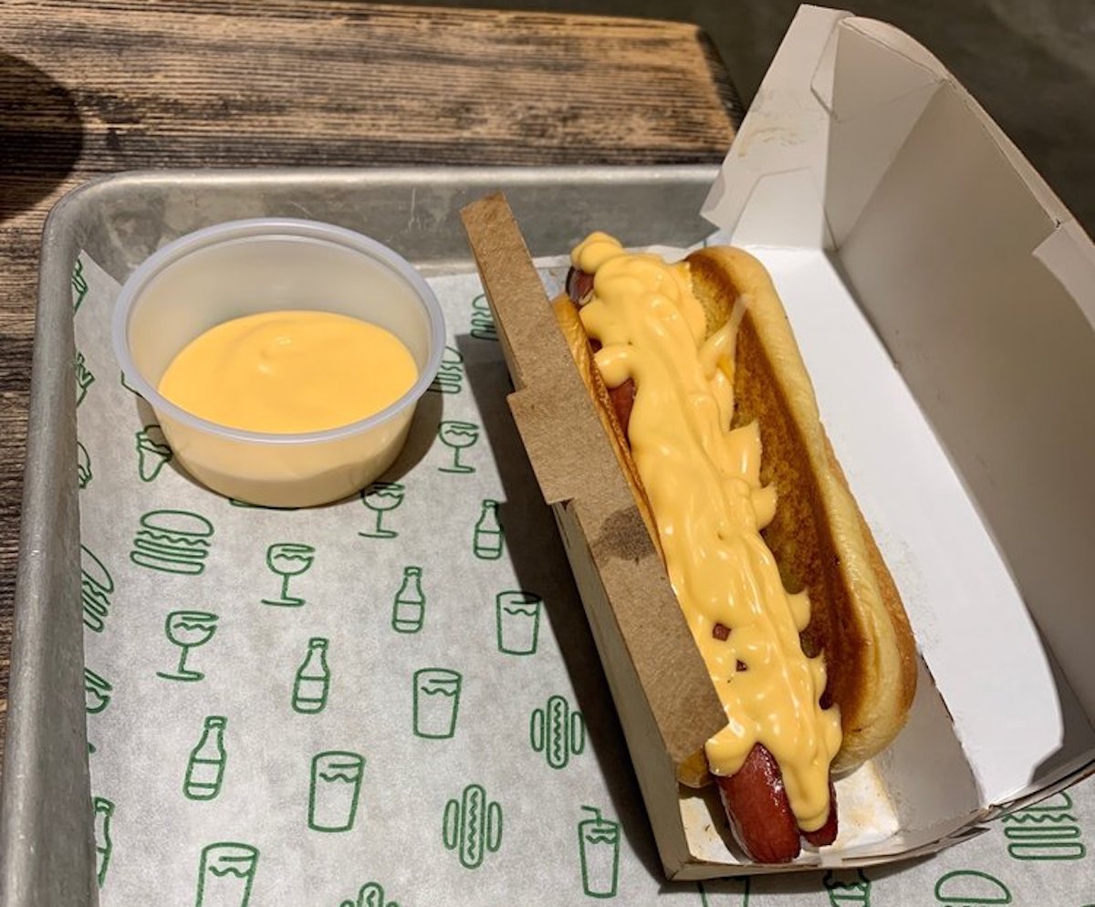 A hot dog topped with melted cheese sits in a cardboard holder next to a small plastic cup of cheese sauce, all placed on a tray with patterned paper featuring food and drink icons.