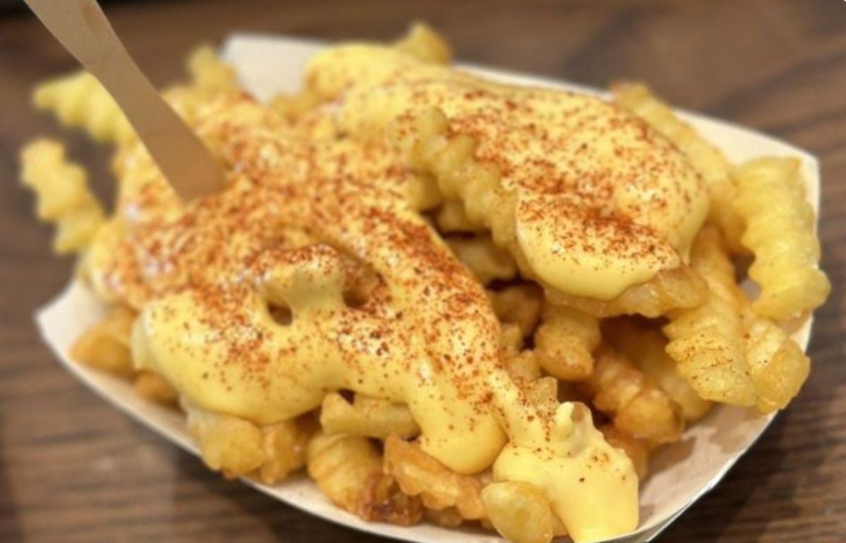 Crinkle-cut French fries in a paper tray, topped with melted cheese sauce and sprinkled with red seasoning, with a wooden fork on the side.