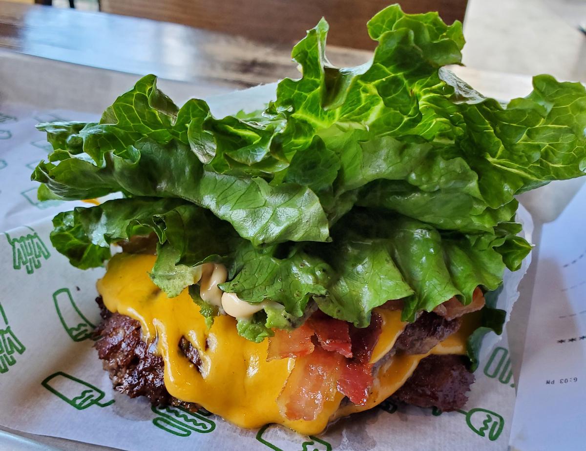 A cheeseburger wrapped in large green lettuce leaves instead of a bun, with melted cheddar cheese, bacon, and sauce visible on a paper-lined tray.