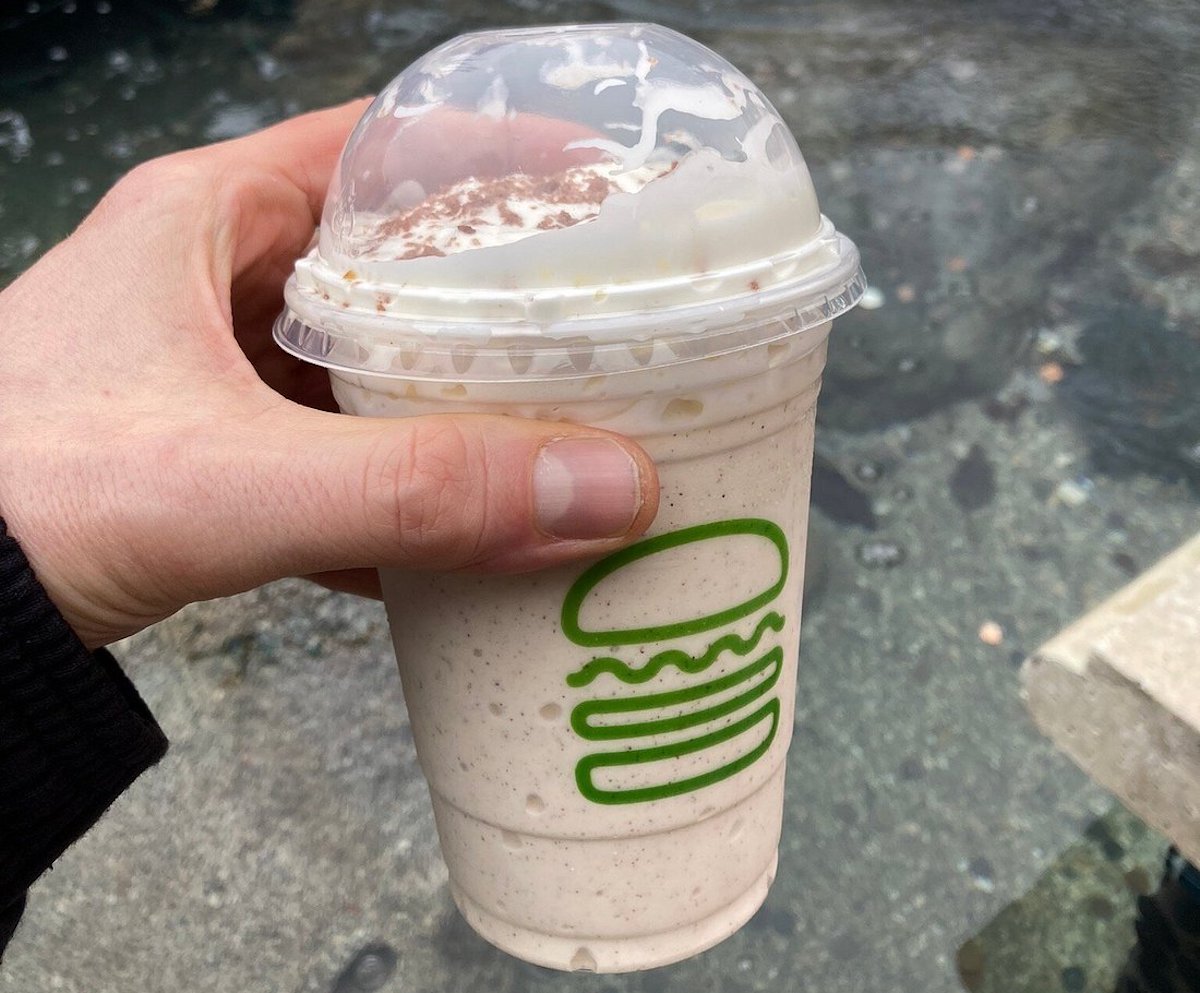 A hand holding a Shake Shack milkshake with whipped cream and a dome lid; the iconic green burger logo is visible on the cup, and the background is outdoors with a stone surface.