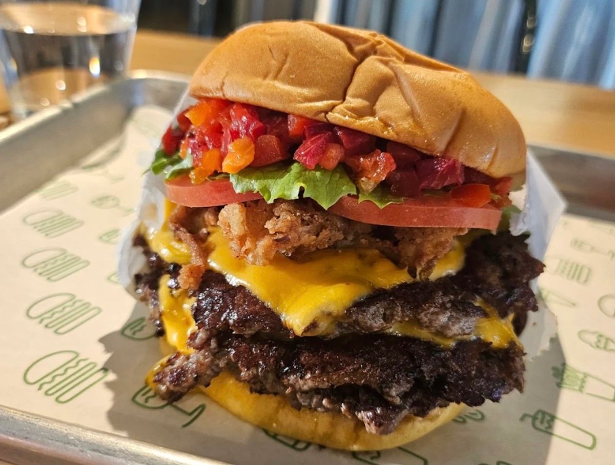A close-up of a cheeseburger with two beef patties, melted cheese, crispy fried onions, lettuce, tomato, chopped red peppers, and a soft bun, served on a metal tray with a cup of water in the background.