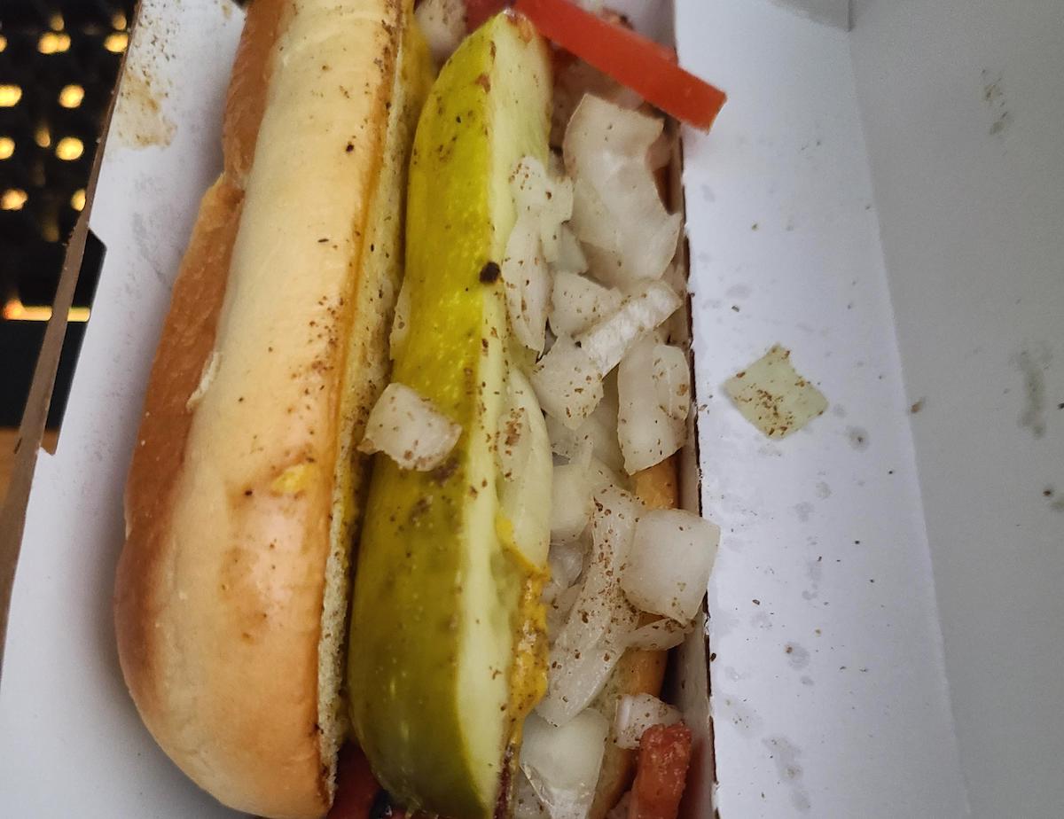 A close-up of a hot dog in a bun topped with chopped onions, two pickle slices, tomato slices, and a sprinkle of black pepper, inside a white cardboard container.
