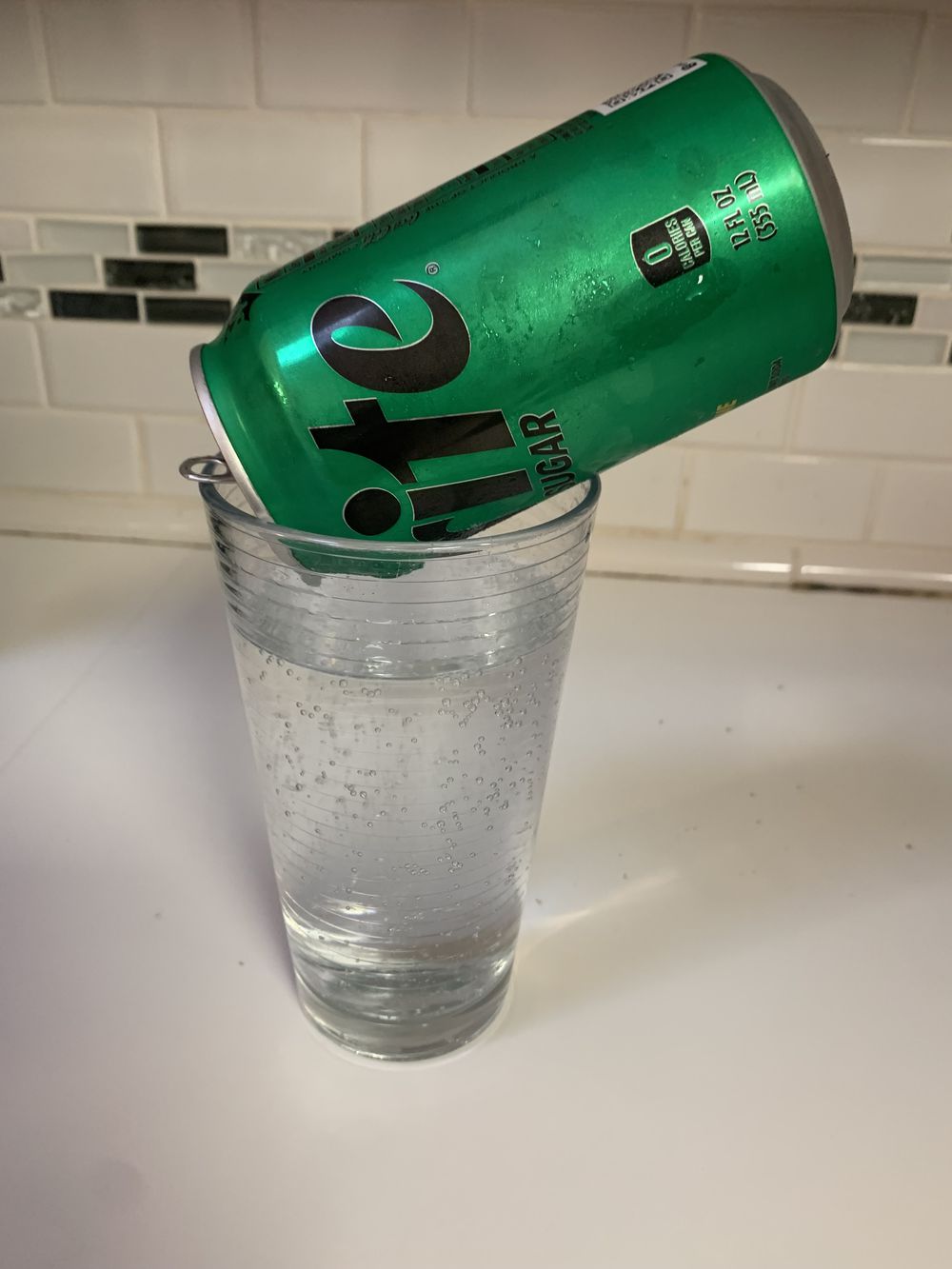 A green Sprite can is propped on top of a clear glass filled with sparkling liquid, possibly Sprite, on a white countertop with a tiled backsplash in the background.