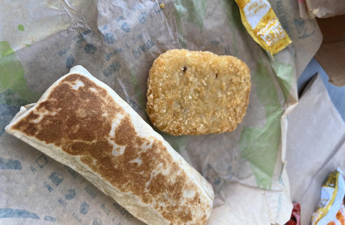 A toasted breakfast burrito and a golden hash brown are placed on a sheet of paper, with a few sauce packets nearby—perfect for anyone seeking the best fast-food breakfast burrito experience.
