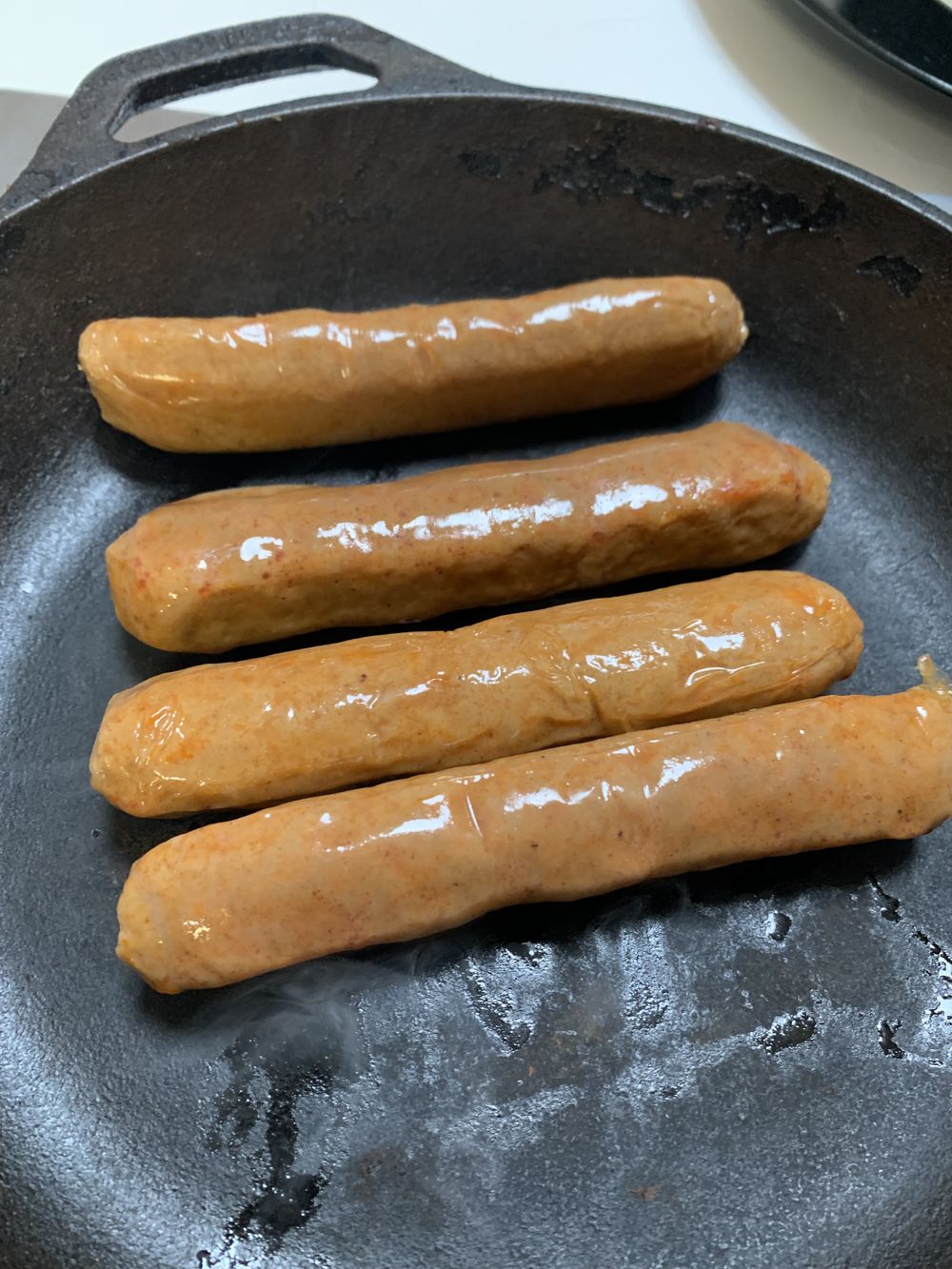 Four uncooked sausages are lined up in a black cast iron skillet, beginning to sizzle and brown slightly as they cook.