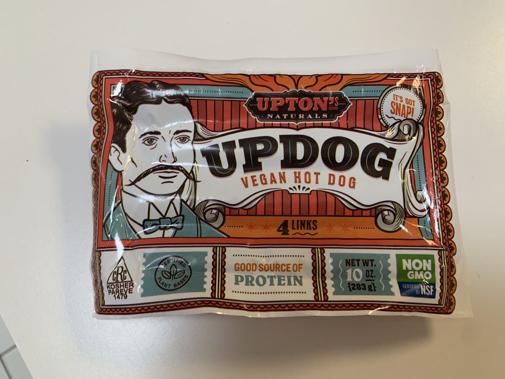 A package of Upton's Naturals Updog Vegan Hot Dog, featuring a vintage-style illustration of a man with a mustache. The label highlights 