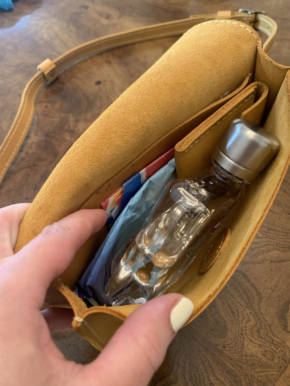 A hand holding open a small brown suede purse reveals a clear reusable water bottle and a pack of tissues inside. The purse is resting on a wooden surface.