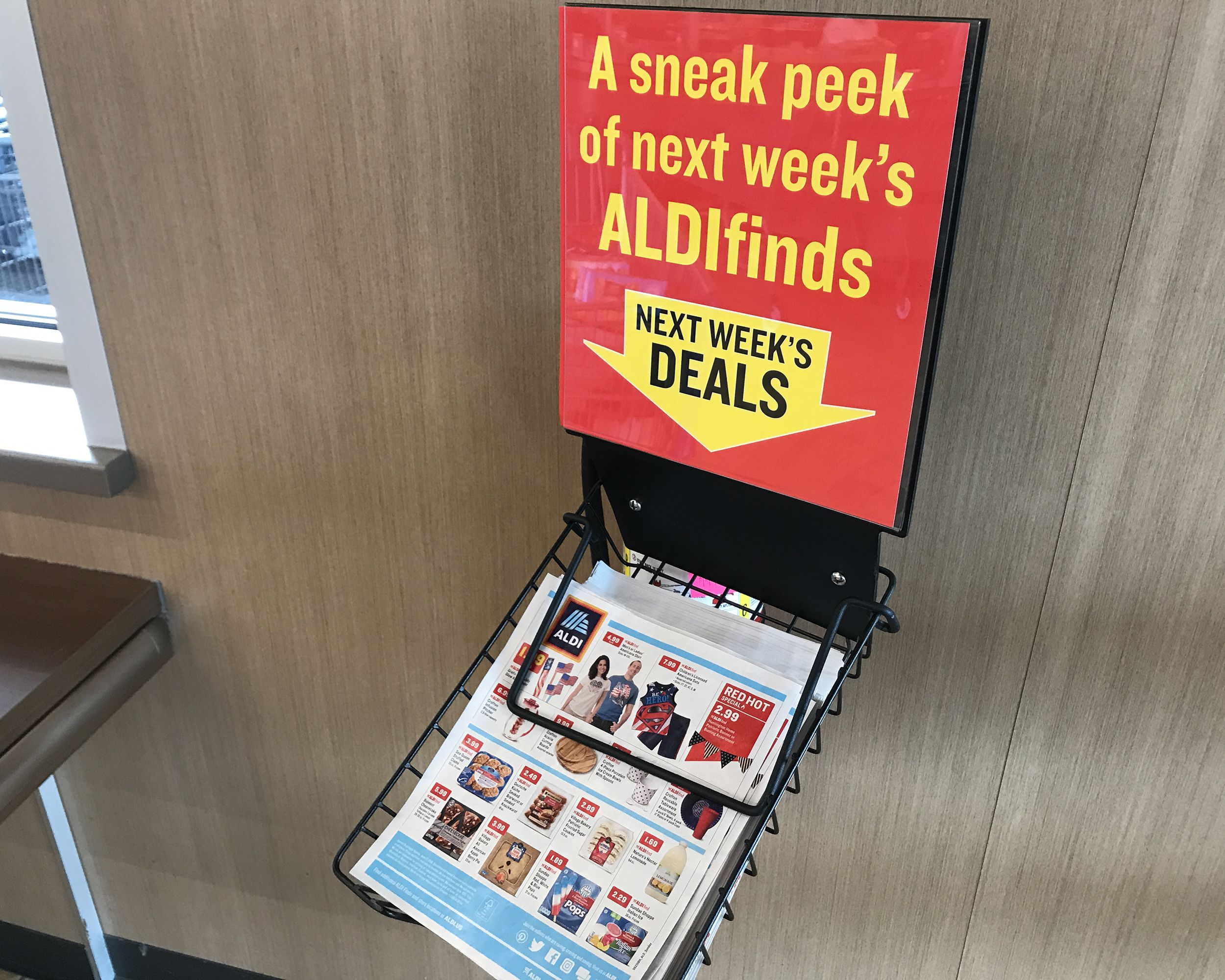 Wire basket holding copies of the AldiFinds deals flyer at an an Aldi store with sign, against wall with window and table in the far left