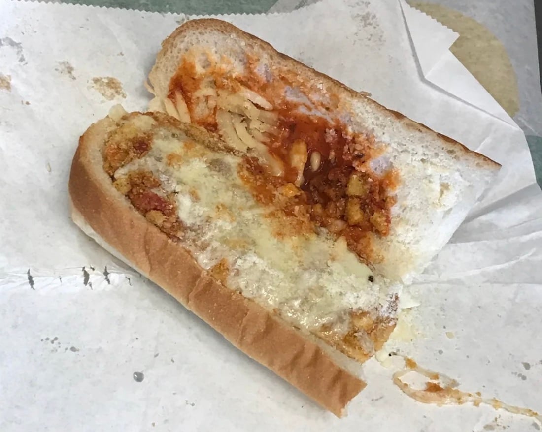 A sub sandwich on white bread is open-faced on white paper, showing melted cheese, tomato sauce, and a few pieces of onion. The filling appears sparse and unevenly spread.