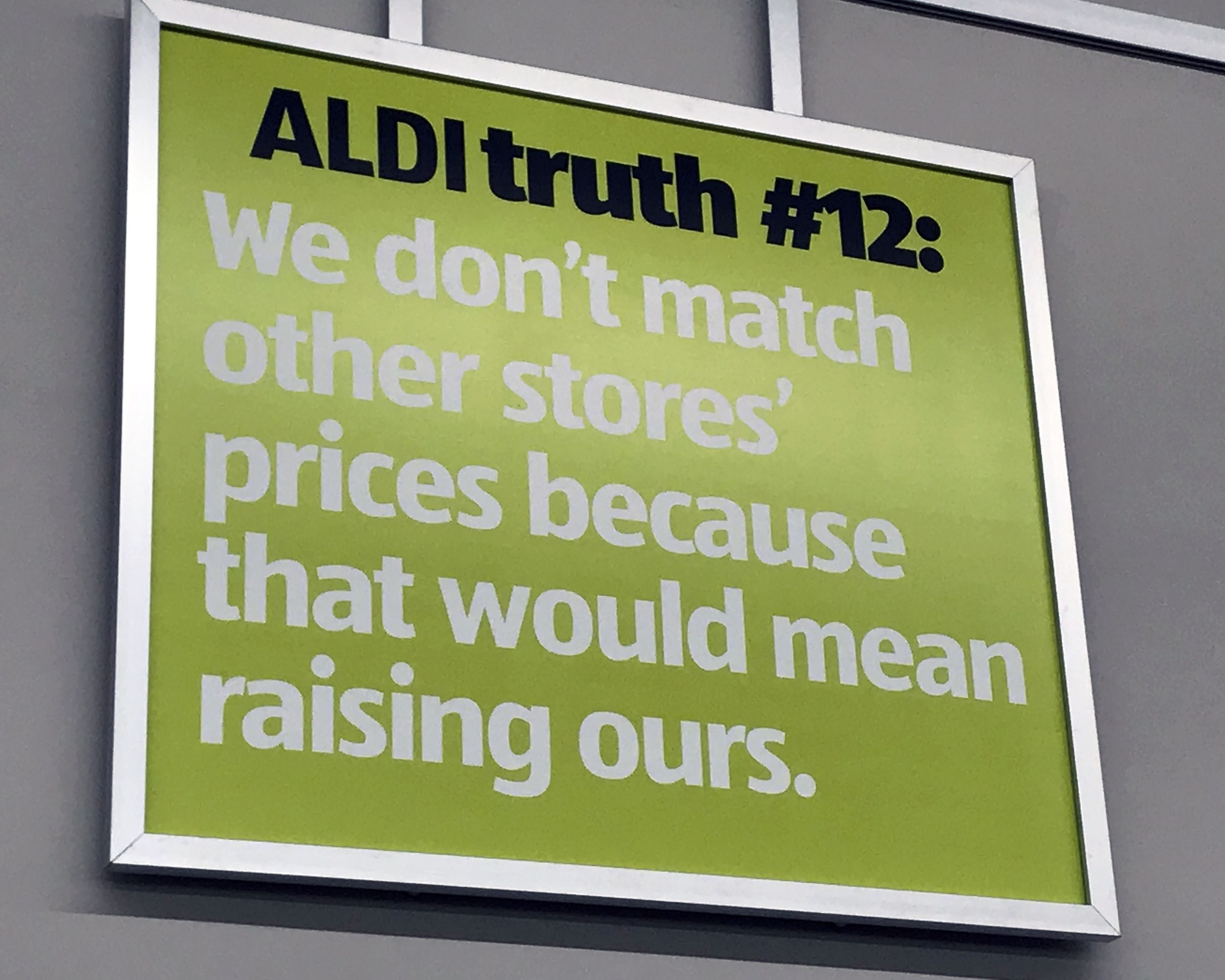 Closeup of Aldi Truth #12 sign at an Aldi store, avocado green, against a wall