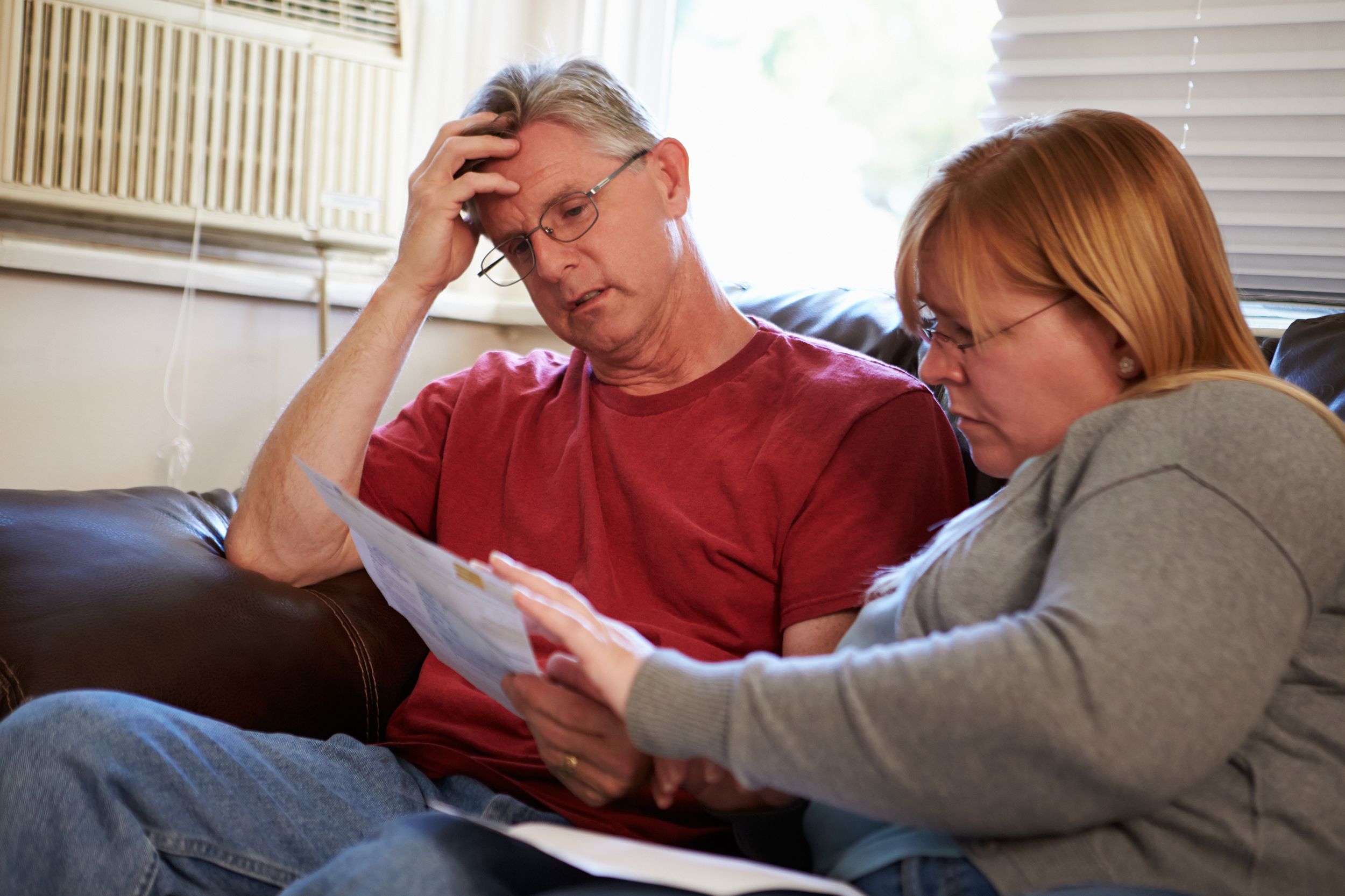 Distressed poor couple looking over bills while seated on a brown leather sofa, air conditioner and window in the background