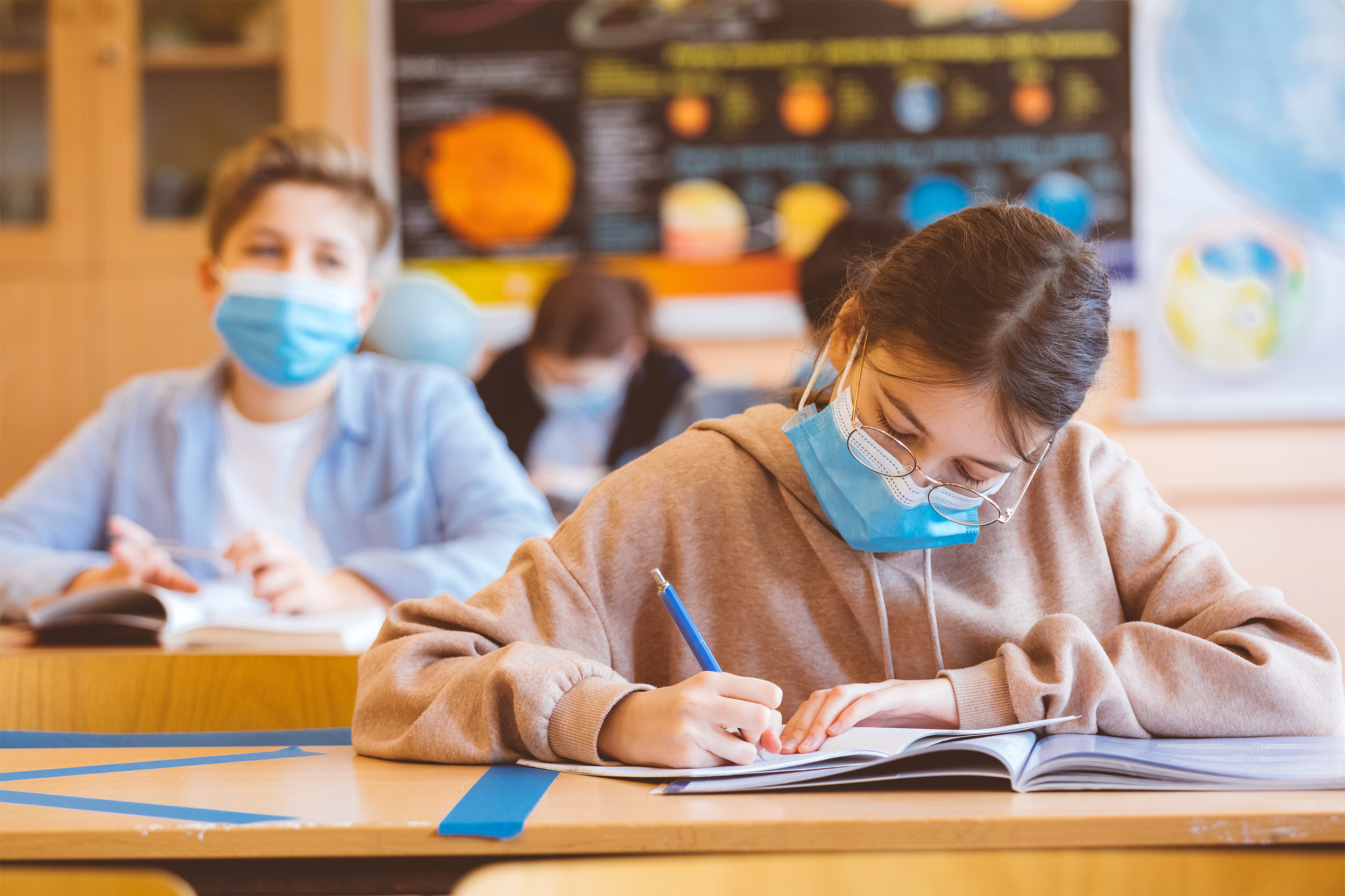 Four middle school children in a classroom wearing face masks while seated at their desks that are social distanced, one girl in the foreground, rest blurred in the background with solar system poster on wall blurred in the background