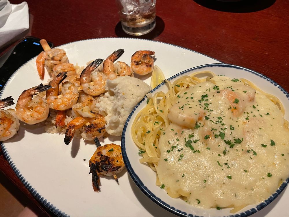 A plate with grilled shrimp skewers, a serving of mashed potatoes, and a bowl of pasta topped with creamy white sauce and parsley. A drink sits in the background on a reddish-brown table.