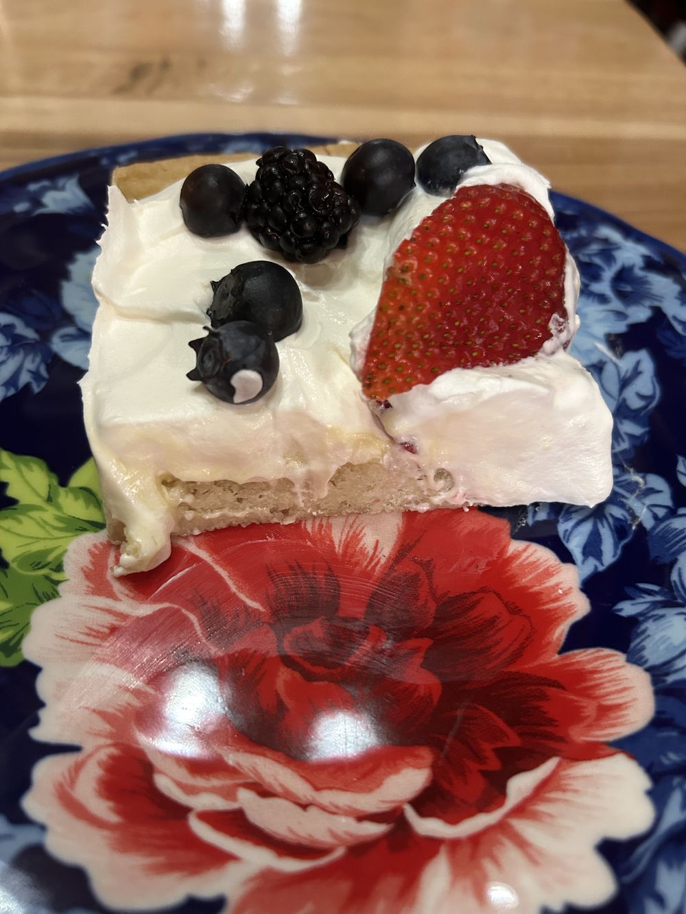 A square slice of dessert with a creamy white topping, garnished with blueberries, a blackberry, and a strawberry slice, sits on a decorative plate with red and green floral patterns.