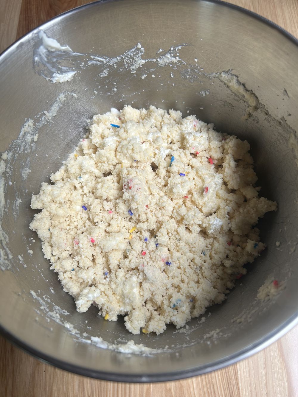 A metal mixing bowl containing coarse, crumbly cookie dough with colorful sprinkles on a wooden surface.
