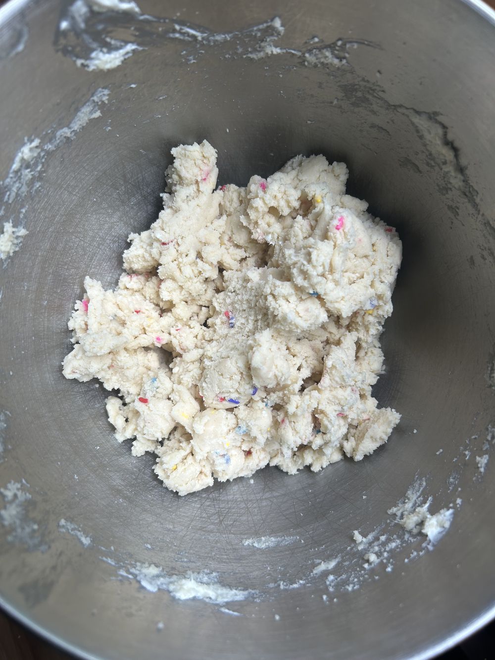 A metal mixing bowl containing a ball of cookie dough with colorful sprinkles mixed in. Some dough and flour residue are visible on the sides of the bowl.