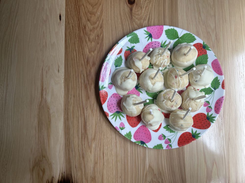 A plate with a strawberry pattern holds a dozen white chocolate-covered cake pops with sprinkles, placed on a light wooden table.