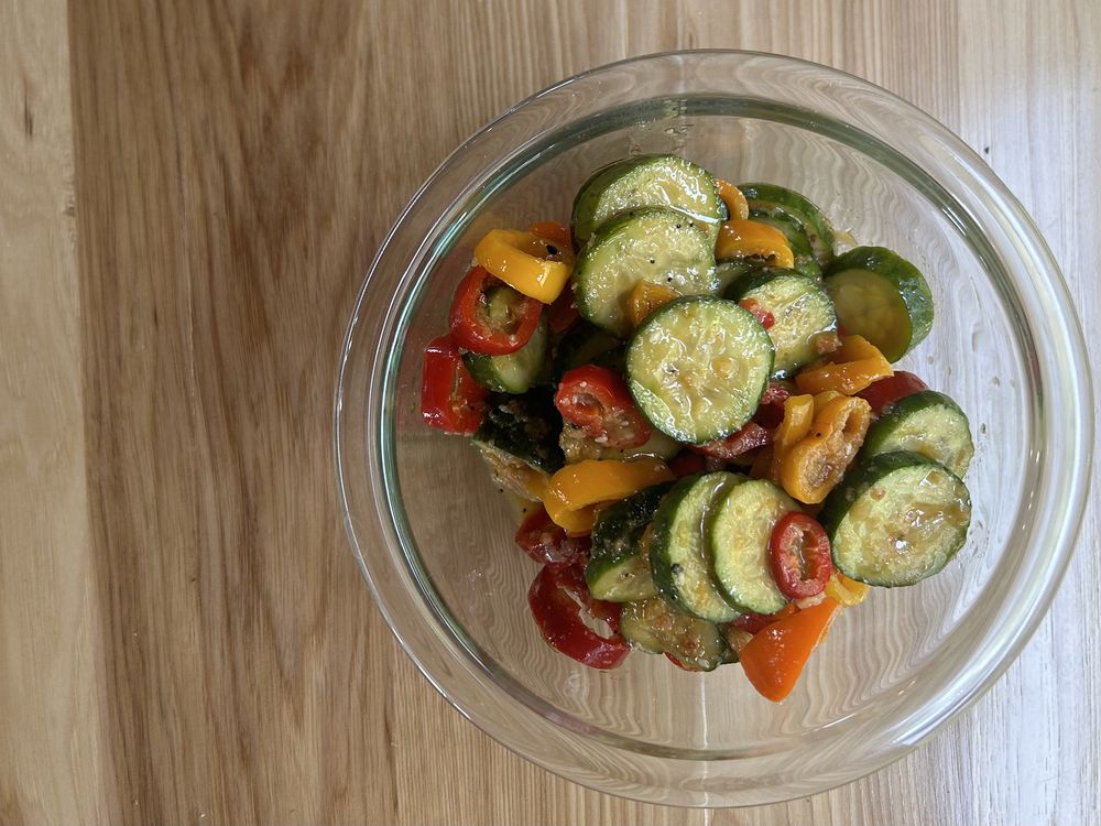 A glass bowl filled with sautéed sliced zucchini and colorful bell peppers sits on a wooden surface.