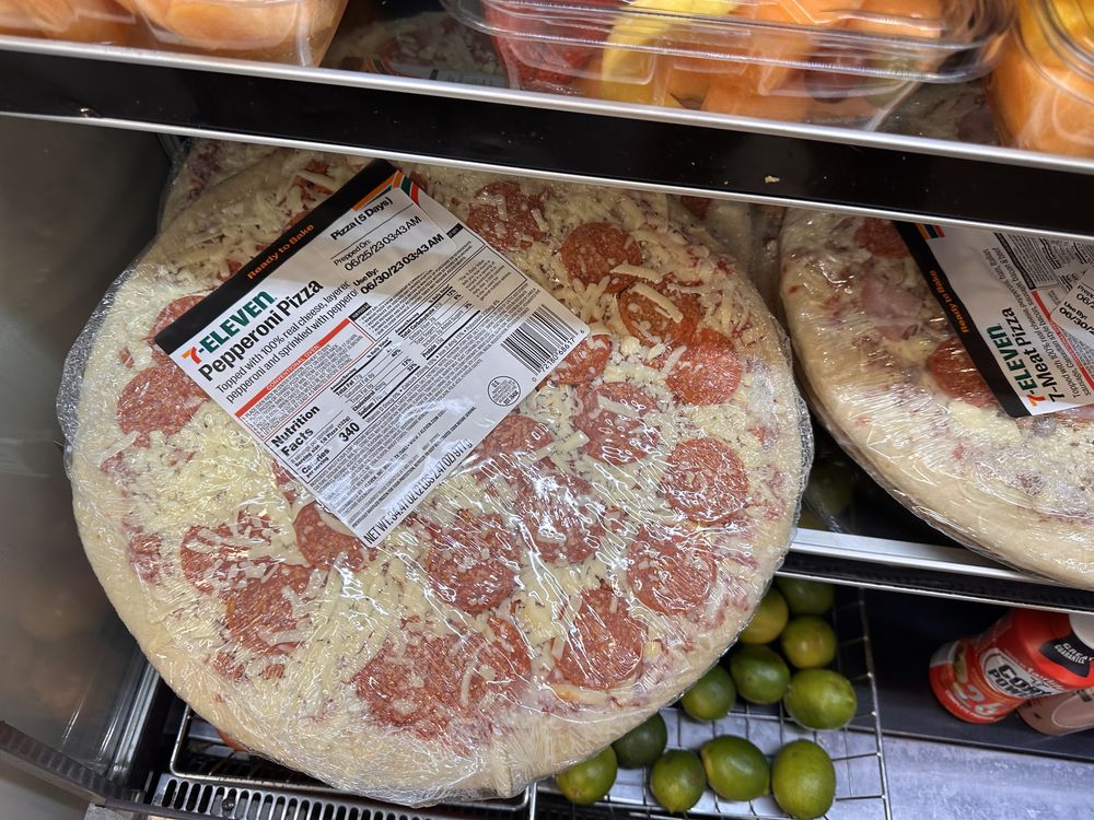 A wrapped 7-Eleven pepperoni pizza sits on a refrigerator shelf above several limes. The pizza label is visible, and other food containers are on the shelf above.
