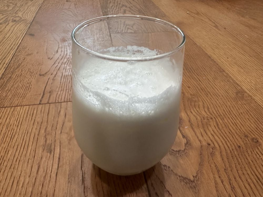 A clear glass filled with frothy white milk stands on a wooden floor with a natural brown grain pattern.