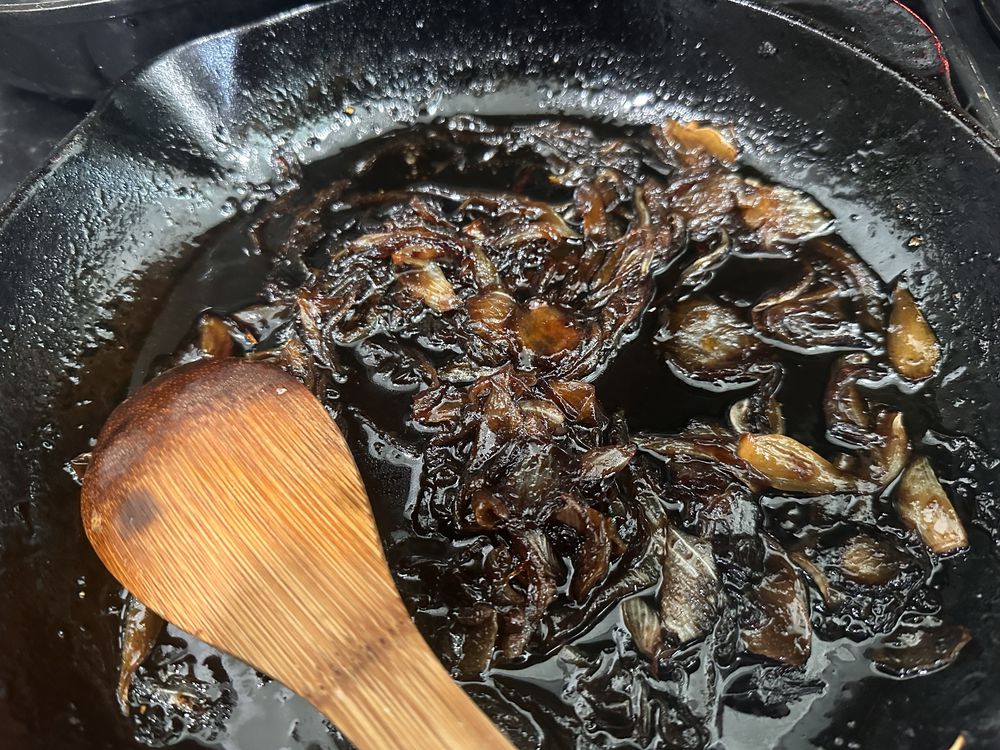 Sliced onions caramelizing in a cast iron skillet, turning deep brown and glossy, with a wooden spoon resting in the pan.