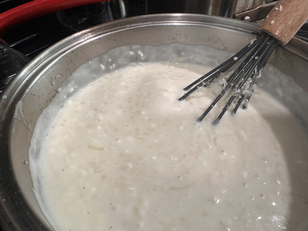 A metal pot on a stovetop filled with thick, creamy white sauce being stirred by a whisk with a wooden handle.
