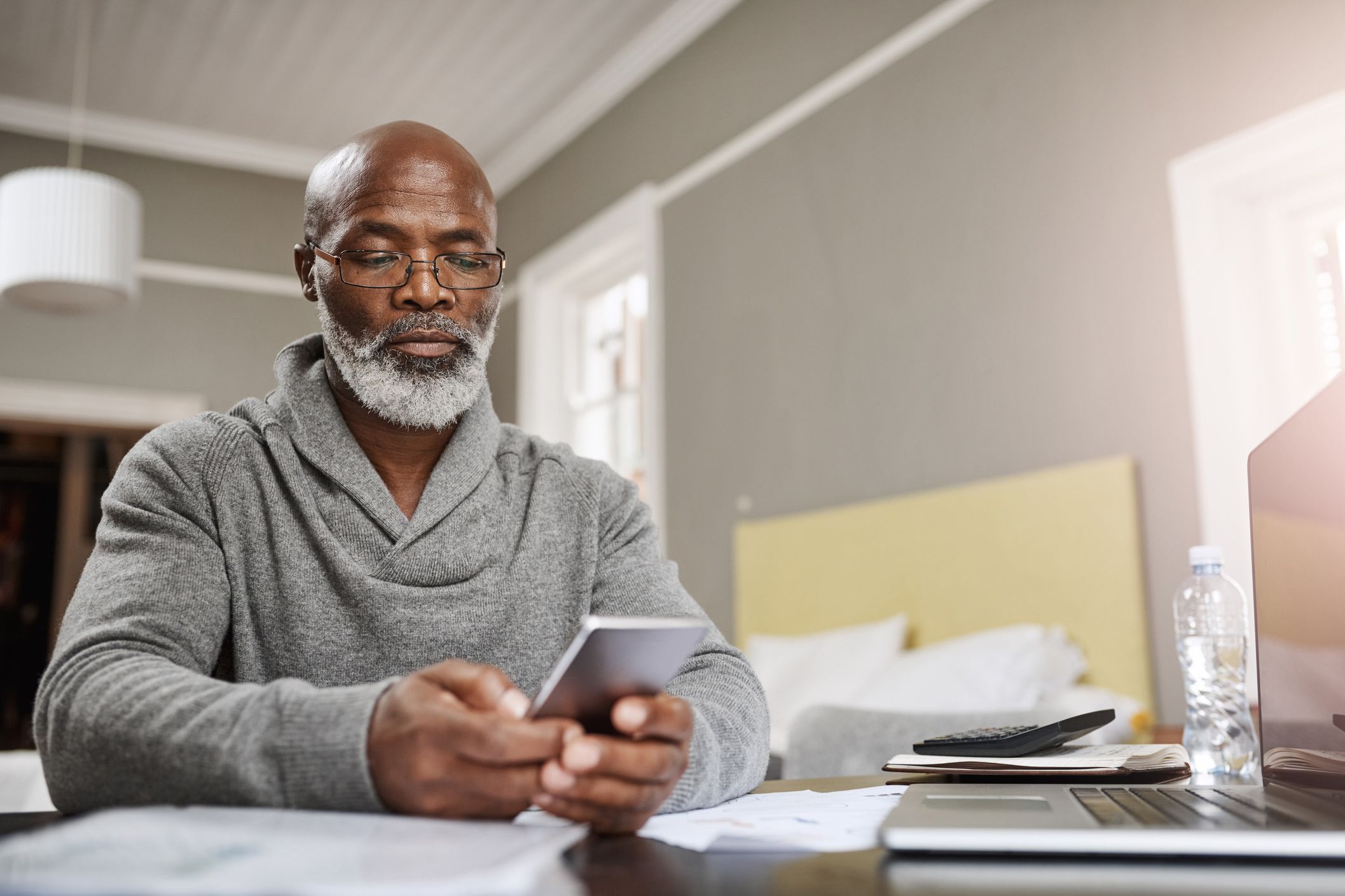 Man looking at smartphone with laptop and tablet to his side on table, hotel bed and room in the background