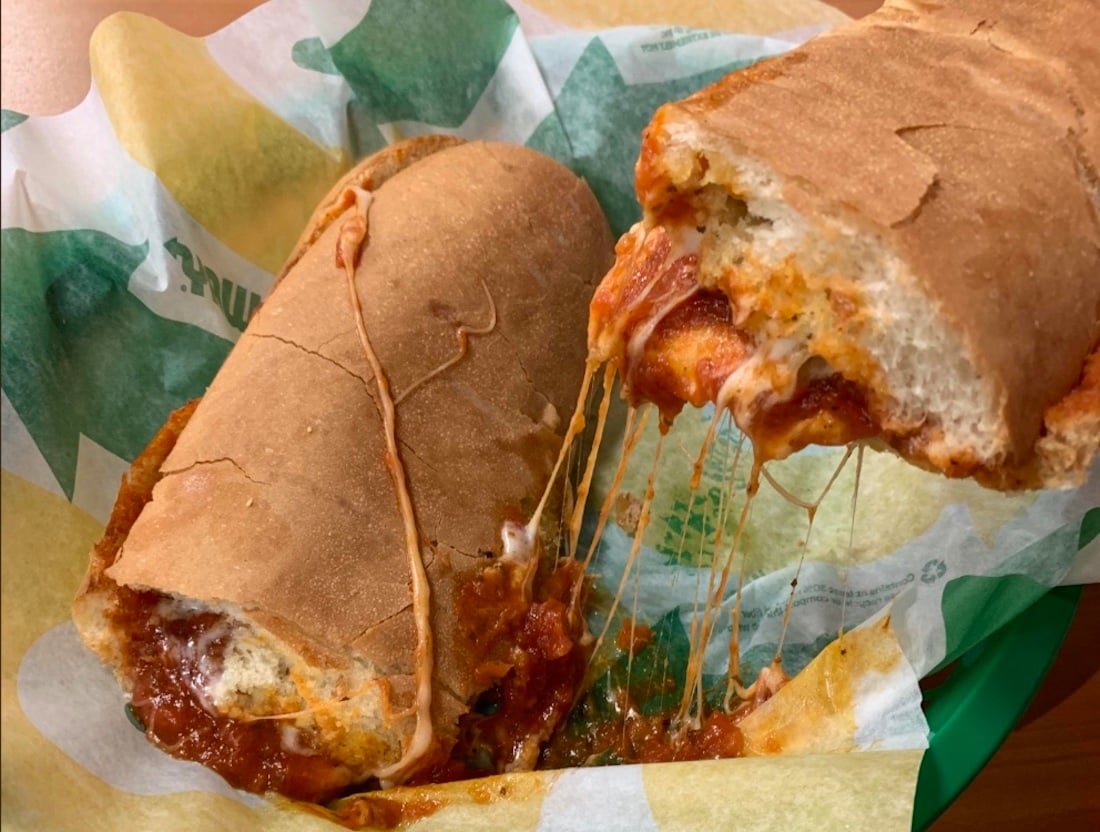 A close-up of a meatball sub sandwich, sliced in half, with melted cheese stretching between the pieces on sandwich paper in a green basket.