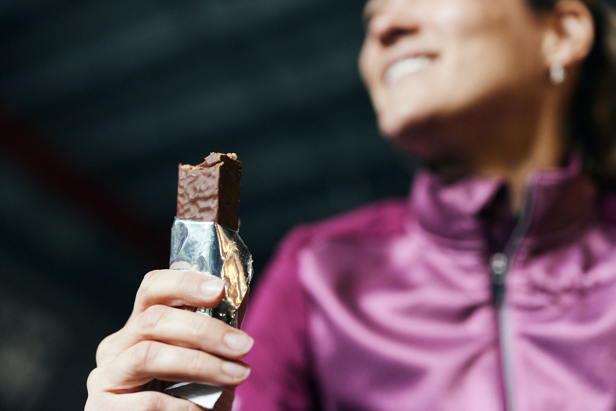 Opened protein bar, with bites out of it, held in the hand of an athletic woman, selective focus, woman is wearing a shiny purple tracksuit jacket and is smiling, blurred
